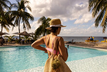 Beautiful Woman With Floppy Hat Wearing Swimsuit Near A Tropical Island Resort Swimming Pool On A Sunny Day During Her Vacation In Paradise. 