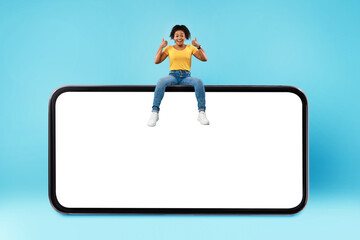 Poster - Positive millennial black woman showing thumb up gesture, sitting on top of huge smartphone over blue background, mockup