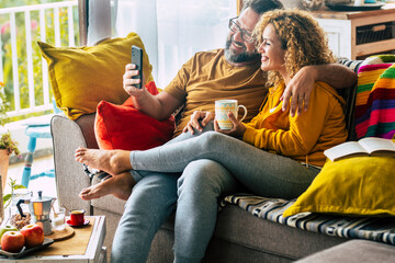 Happy adult couple at home doing video call with friends sitting on the sofa during morning breakfast and relax together - man and woman with connection mobile technology enjoying love 