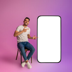 Poster - Positive young Arab guy sitting on chair, pointing at huge smartphone with empty screen in neon light. Mockup