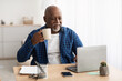 Black Mature Man Working On Laptop Drinking Coffee At Workplace
