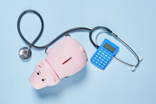 Piggy Bank With Stethoscope And Calculator On Blue Background, Concept Of Collecting Money For Health