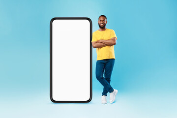 Poster - Full length of funky young black guy leaning on huge blank smartphone on blue background, mockup for mobile app