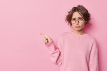 Displeased European Woman With Bob Hairstyle Has Sulking Look Points Away At Blank Copy Space Shows Place For Your Advertisement Wears Round Spectacles And Sweater Isolated On Pink Studio Wall
