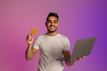 Poster - Portrait of cheery millennial Arab man holding credit card and laptop in neon light. Online shopping, remote banking