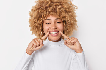 Wall Mural - Look at my perfect orthodontic smile. Positive woman with curly bushy hair cheerful expression points at white teeth smiles broadly takes care about dental health poses indoor expresses happy emotions