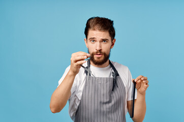 Wall Mural - emotional hairdresser in gray apron with scissors and comb on blue background trendy hairstyle