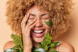 Beauty and skin care concept. Overjoyed woman laughs out happily covers face with hand smiles toothily has natural clean skin applies green collagen patches under eyes for reducing wrinkles.