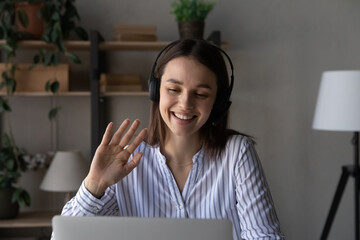 Happy young student girl in headphones with mic talking on video call to teacher, tutor, waving hello at webcam, looking at screen, smiling, laughing, speaking. Online virtual training class concept