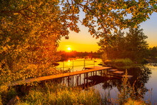 Sunset Over The River. Wooden Village Bridge. Clear Sky. Smooth Surface Of Water. View From The Shore Through The Foliage. Russia, Europe.