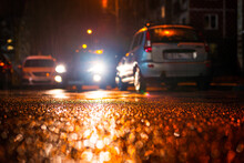 Night City In The Rain. Headlights Of A Driving Car. City Courtyard. Parked Cars. Residential Buildings. Falling Raindrops. Colorful Colors. Focus On The Asphalt. Close Up View From The Asphalt Level.