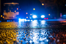 Night City In The Rain.  Headlights Of Approaching Cars. Parked Bus. Falling Drops Of Rain. Colorful Colors. Focus On The Asphalt. Close Up View From The Asphalt Level.