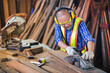 © pornchai - Asian elderly carpenter In work clothes with work safety equipment such as gloves,glasses, ear-muffs or noise-cancel equipment are using an electric planer to smooth out the wood to produce furniture