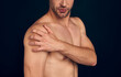 Handsome young bearded man isolated. Portrait of naked muscular man is standing on dark blue background. Man holding his shoulder. Experiencing shoulder pain