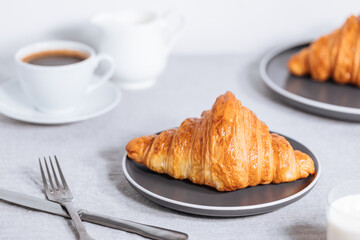 Croissant in bright white environment, with cup of coffee and milk and other croissant in blurry background. Simple and elegant breakfast setting and scene.