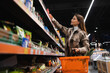 © Andrii  - Pretty woman with shopping basket looking at groceries on supermarket shelves
