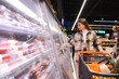 © Andrii  - Pretty young woman with shopping trolley in supermarket taking groceries from refrigerator