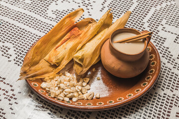 Sweet pineapple and blue tamales and red chili tamales, accompanied by white masa atole. Typical Mexican food. On a clay plate and accompanied by corn and cinnamon.