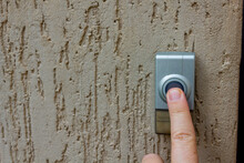 Selective Focus On A Caucasian Finger Pressing A  Round Button On An Outdoor Doorbell. The Doorbell Is Mounted On A Textured Wall