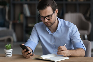 Poster - Focused confident businessman in glasses holding smartphone, taking notes, writing down important information in notebook, sitting at desk, entrepreneur planning workday, student watching webinar