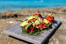Fresh Avocado And Tuna Fish On Mexican Tostada For Lunch. Food Is On A Wooden Board On A Beach With Tropical Turquoise Ocean Water In The Background.