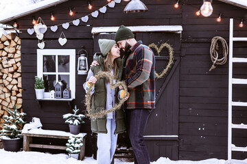 Wall Mural - young couple in love beautiful Asian girl and bearded man in stylish winter clothes standing near porch of country house with handmade wreath of dried grass in shape heart, winter spirit valentine day