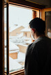 Young man with mask in quarantine looking at the snowfall through the window of his house
