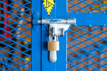 A Key Padlock Is Locking On Metal Fence Gate Knob Of The Hand Tool Storage Box (as Blurred Backgroung). Industrial Object Photo. Close-up And Selective Focus.