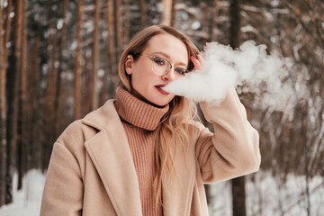 Blonde mid age woman smoking vape or cigarette in winter forest. Girl in coat blowing smoke outdoors.