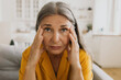 © shurkin_son - Portrait of woman suffering from stress and headache, sitting on couch massaging temples with fingers, looking at camera with painful face expression, isolated against home background