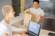 © rh2010 - Blonde woman choosing groceries in online store on laptop. Partial of man with shopping bags on background of girl. Concept of online shopping and e-commerce. Idea of healthy eating. Kitchen interior