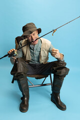 Sticker - Portrait of young man, fisherman with fishing rod, spinning and equipment sitting isolated over blue studio background