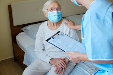 Wall Mural - Hospital nurse wearing a face mask, holding a clipboard with medical history for an elderly lady. Senior woman and her designated care giver discussing test results. Background, close up, copy space.