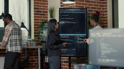 Software engineer holding digital tablet analyzing code on wall screen tv explaining errors to colleague programer in it startup office. Team of coders collaborating on ai innovation group project.