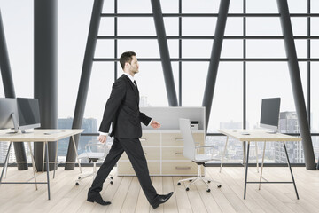 Poster - Attractive european businessman walking in modern concrete coworking office room interior with panoramic window and city view, wooden parquet flooring, equipment, daylight and furniture.