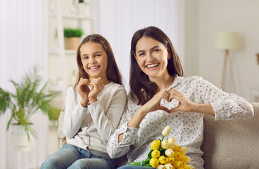 Happy loving mom and child celebrating Mother's Day together. Portrait of happy Caucasian mother and daughter sitting on sofa, looking at camera, smiling and doing heart shape gesture with hands