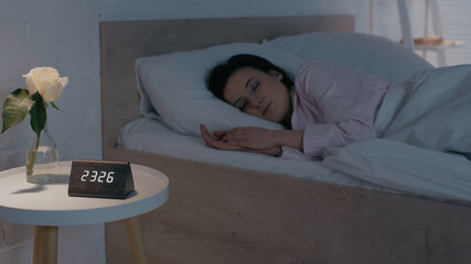 Flower and clock on bedside table near woman sleeping at home.