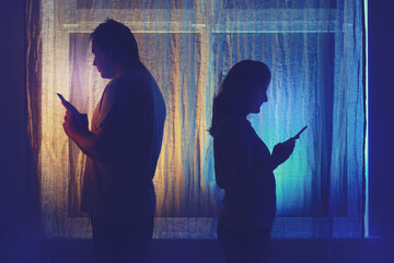 Wall Mural - Silhouette of a man and a woman with phones at the night window. Couple husband and wife having relationship problems in the evening light of the home living room