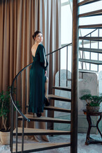 Beautiful Caucasian Woman Wearind Green Long Evening Dress Posing At Camera Standing On Spiral Staircase In Beautiful Apartment. Elegant Slim Pretty Lady In A Dress With A Deep Cut On The Back