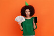 © khosrork - Shocked woman with Afro hairstyle wearing green casual style sweater standing with open mouth, holding dollar banknotes and phone with blank screen. Indoor studio shot isolated on orange background.