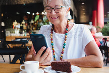Portrait Of Senior Smiling Woman Relaxing For A Break At Coffee Shop With Chocolate Cake And Coffee Cup. Carefree Elderly Woman With Eyeglasses And Necklace Using Mobile Phone
