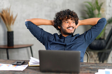 Unshaved man sitting at the table and smiling, while feeling comfort and relaxation at his workplace. Confident office worker sitting in front of the laptop and looking away