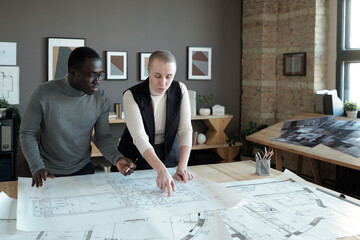 Poster - Two confident engineers in smart casualwear discussing sketch on blueprint while standing by table in large office