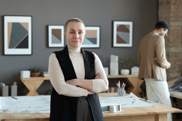 Poster - Successful female architect crossing arms by chest while standing by table with blueprints on background of young man