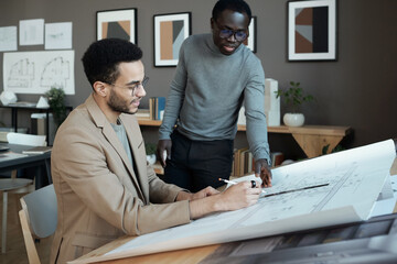 Poster - Two young intercultural businessmen discussing sketch of architecture on blueprint while one of them drawing lines