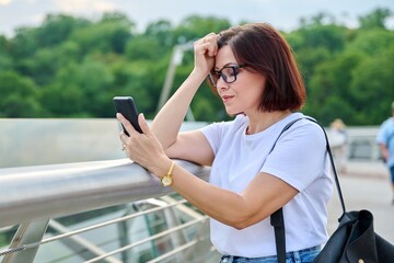 Wall Mural - Middle-aged woman using smartphone, walking in the summer city