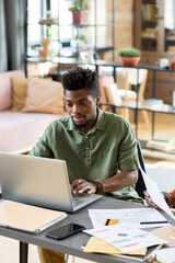 Poster - Young businessman of African ethnicity looking at laptop display while networking by table in home environment