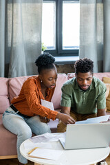 Poster - Young African couple in casualwear discussing financial papers or tax payment form while sitting on sofa in front of laptop