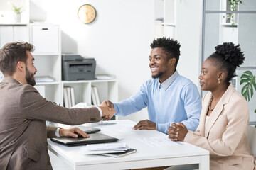 Poster - Young elegant lawyer in formalwear congratulating African couple while shaking hand of happy man over table in office