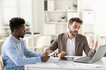 Poster - Confident businessman making presentation of online data on laptop display to young African colleague at working meeting in office